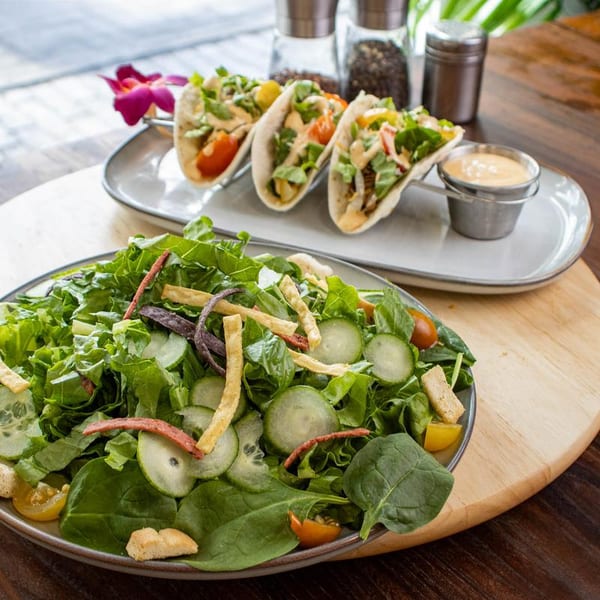 A fresh green salad with cucumbers and tomatoes, alongside three tacos with sauce and a purple orchid.
