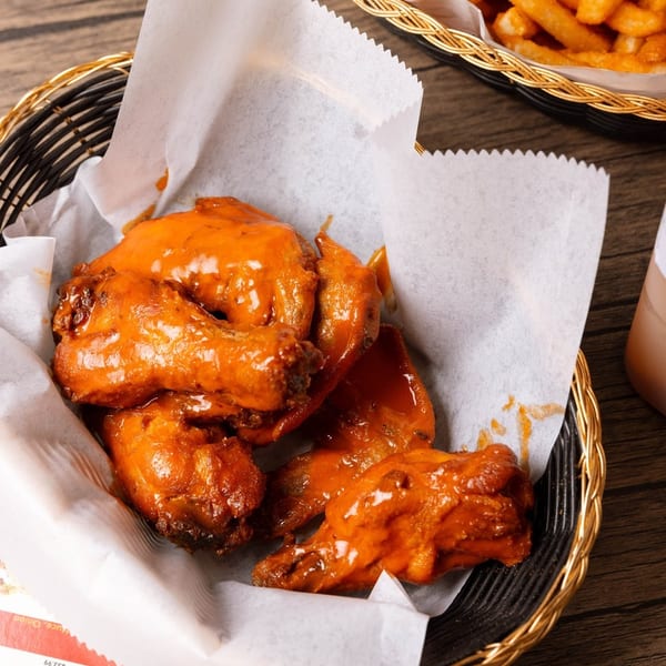 A basket of buffalo chicken wings and a basket of french fries on a wooden table.